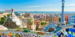 View of Park Güell in Barcelona, Spain, showing colorful mosaic tiles in the foreground and panoramic cityscape with the Mediterranean Sea in the distance under a bright blue sky.